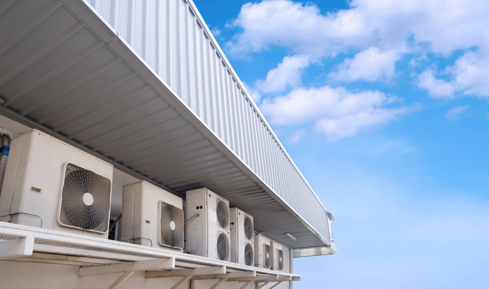 Row of air conditioner compressors under steel roof eaves of convenience store against blue sky